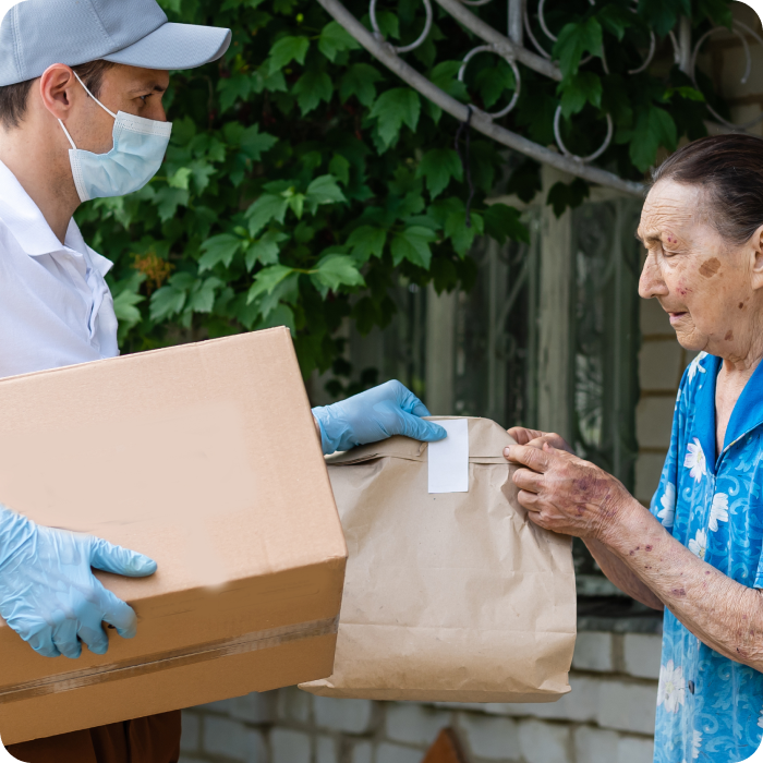 A delivery person wearing a cap, face mask, and gloves hands a package and a paper bag to an elderly woman outside a building. The woman, dressed in a blue floral shirt, reaches for the items with one hand while holding the door with the other. Green foliage frames this moment of trust and connection.